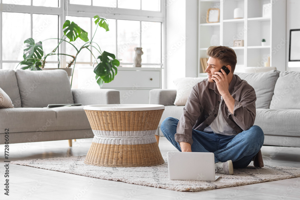 Young man with laptop talking by mobile phone at home