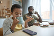 Portrait of African American boy eating breakfast at kitchen table with family and looking at smartphone screen watching videos online