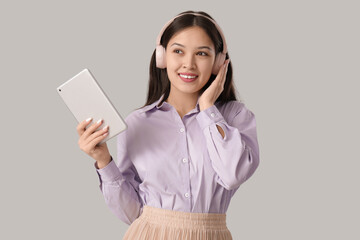 Canvas Print - Beautiful young Asian woman in headphones listening to audiobook with modern tablet computer on grey background