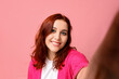 Smiling young redhead woman taking selfie on pink background