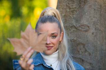 Pretty young blonde woman with a ponytail in her hair covers one eye with a dry leaf. Focus on the face. Autumn and winter concept.