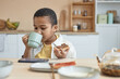 Front view portrait of young boy watching videos via smartphone while eating breakfast at kitchen table