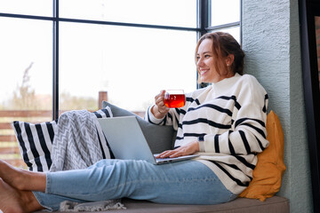 Wall Mural - Young woman using a laptop computer while relaxing on the windowsill sofa at home