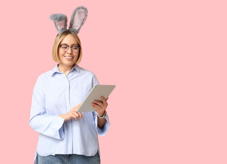 Wall Mural - Happy adult businesswoman in bunny ears with tablet computer on pink background. Easter celebration