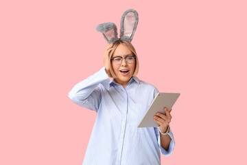 Wall Mural - Shocked adult businesswoman in bunny ears with tablet computer on pink background. Easter celebration