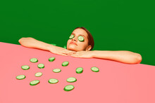 Spa and relaxation. Young woman peeking over table with cucumber slices on face, homemade refreshing mask against green background. Concept of food pop art photography, creativity, quirky style