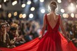 A model girl in a red long dress, with her hair styled, walks along the floor under the spotlight. Spectators on both sides watch the fashion show. Back view
