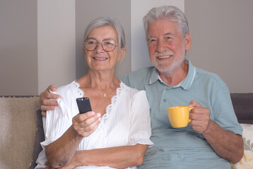 Wall Mural - Happy bonding senior retired couple sitting on sofa watching television, enjoying peaceful and funny moments together at home.