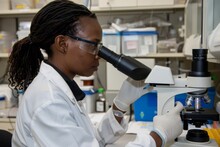 A Black Woman Wearing Glasses And Lab Coat Is Looking Through The Lens Of An Microscope At Samples On A Slide. She's Sitting Inside A Laboratory With Shelves Filled With Various Vials And Test Tubes.