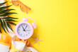 Morning summer vibes: Top view photo of colorful and energetic start with an alarm clock, sunscreen, and beach essentials on a lively yellow background, signaling vacation time