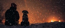 Person And A Dog Siting Outside At Night In Winter When The Snow Is Snowing. 