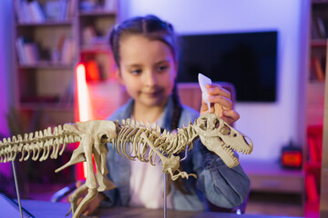 Small Caucasian girl scientist six years old in casual clothes sitting at table examine skeleton of dinosaur. Happy preschool child gluing bones making model of tyrannosaurus at evening home.