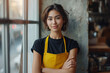 Young female entrepreneur with yellow apron and arms crossed in cozy cafe interior