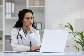 A professional Hispanic female doctor is intently working on her laptop in a modern clinic office, displaying dedication and care.