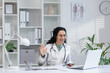 Friendly Hispanic female doctor in white coat waving during a video call consultation in a bright office setting.