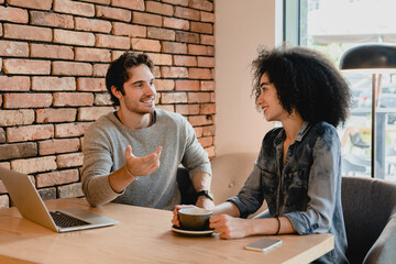 Two young colleagues using laptop while drinking coffee in cafeteria. Coworkers customers couple having conversation, doing freelance job projects online in cafe restaurant