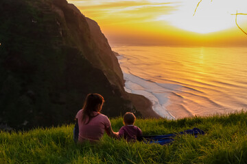 Wall Mural - Mother with child on meadow watching breathtaking sunset at viewing point Miradouro do Ponta da Ladeira, Madeira island, Portugal, Europe. Panoramic view of majestic coastline of Atlantic Ocean. Awe