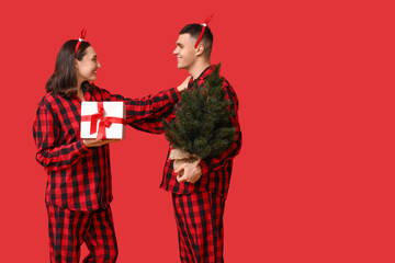 Poster - Happy young couple in pajamas, with Christmas tree and gift on red background