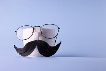 Wall Mural - Man's face made of artificial mustache, glasses and cup on light blue background. Space for text