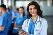 © Kate Mayer - Young smiling female doctor holding digital tablet in conference room, doctors in blue uniform sitting at conference table, depth of field image. Generative AI