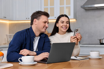 Wall Mural - Happy couple with gadgets shopping online at wooden table in kitchen