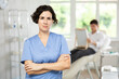 © JackF - Positive middle-aged woman doctor posing against background of doctor's cabinet with patient waiting for consultation