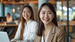 © Surachetsh - two young Asian women smiling and sitting in front of their laptops, with a modern office space as the background.