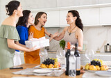 Fototapeta Paryż - Girls hug and kiss on cheek when they meet, company group of three guests gives gift to birthday woman. Treats, food, alcohol and snacks on table in background. Concept of home party