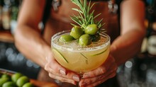   A Close-up Photo Of A Person Sipping From A Drink In A Wine Glass, Topped With Green Olives And Fresh Sprigs Of Rosemary