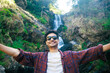 Stylish young traveler or vlogger holding camera and shooting himself, recording video report of his journey for his vlog, standing at Iruppu waterfalls in Coorg or Madikeri, Karnataka, India. 