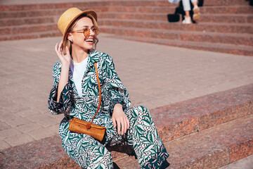 Young beautiful smiling blond hipster woman in trendy summer hippie costume clothes. Carefree female posing in the street at sunny day. Positive model outdoors at sunset. Cheerful and happy in hat