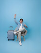 Happy joyful young Asian tourist man listening to music, raising his arm dancing sitting on chair with luggage in airport going to travel on summer holiday isolated on blue background.