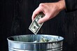 A hand is holding a dollar bill and throwing it into a trash can. Concept of wastefulness and disregard for money