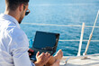 Man working on yacht by sea with laptop. Traveling on sailboat. Traveler using computer, Internet. Freelancer workplace and office in vacation. Successful business lifestyle. Close-up of blank screen