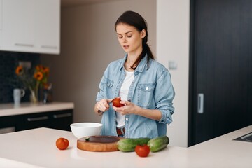 Wall Mural - Beautiful Caucasian female cook preparing healthy salad in the kitchen, surrounded by fresh organic vegetables and cutting fruits, displaying a confident and focused expression.