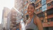 A beautiful young African-American woman in sports clothes smiles, holding up a bottle of water after a workout on the street, against the background of sun-drenched buildings