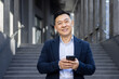 © Tetiana - Close-up portrait of a young Asian man in a suit standing outside an office building, holding a phone and smiling at the camera