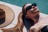 Fototapeta Na sufit - Beauty fashion model girl lying near pool and relaxing, sunbathing. Beautiful young woman portrait over pool background, sun tan concept. Vacation