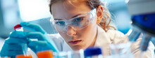 A Woman Wearing A Lab Coat And Goggles Is Looking At A Collection Of Test Tubes