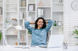 A relaxed Hispanic woman takes a break while working remotely in her bright home office, embodying work-life balance.