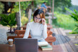 © Wasana - Young woman wearing glasses sitting at a garden table, looking thoughtful with a laptop and notebooks in front of her.