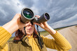 female ornithologist birdman watches birds with binoculars on dirt gravel road against a background of a stormy sky