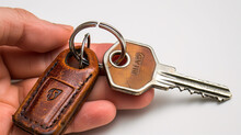 Close-up Of A Hand Holding A Key With A Leather Keychain On A White Background.