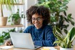 An attractive black woman of 30 years old, wearing a blue sweater and glasses, smiling and looking happy, sitting at a table with a laptop surrounded by plants, and working in a home office