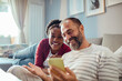 © Davor - Joyful interracial couple looking at smartphone and laughing together on sofa