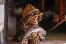 Old Figurine Of Trolls, Holding A Fish In Hands On A Blurry Background