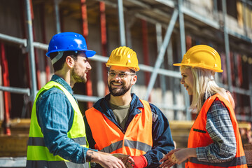 A group of three workers at a construction site wearing hard hats, safety glasses and reflective clothing, smiling and conversing.