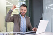 © Liubomir - A triumphant businessman raises his fist in a gesture of success while working on his laptop in a modern office setting.