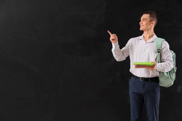 Wall Mural - Male student with book and copybooks pointing at something on blackboard background. End of school concept