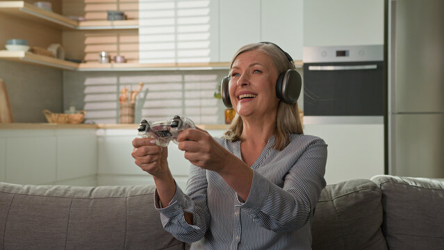 Funny smiling mature caucasian woman playing video game console in headphones using joystick controller at home on couch elderly female play computer games older people and modern technology concept